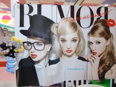 Balloon Seph next to a purikura (photobooth) that says "Rumors are rampant!" and shows three glamorous blonde girls