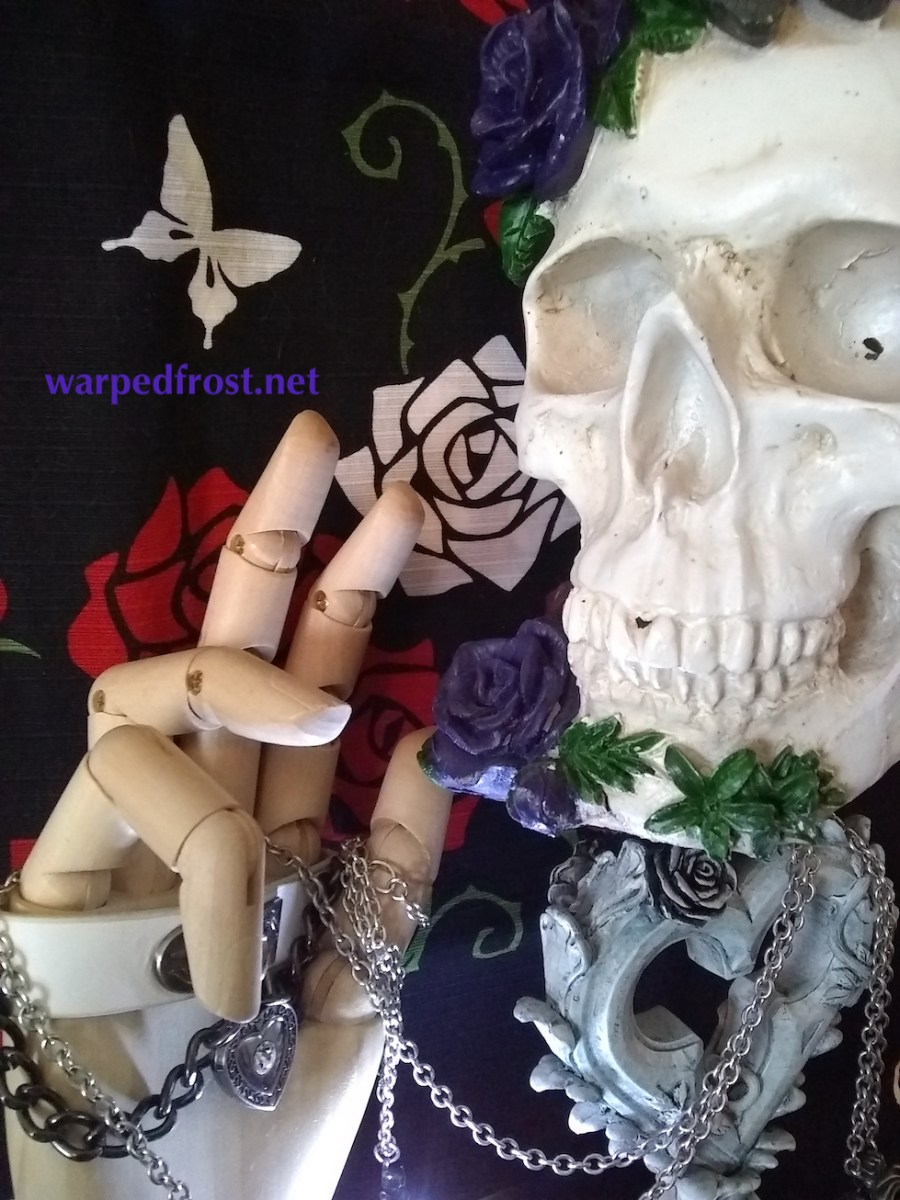 A still life arrangement showing a skull with flowers on it, a wooden mannikin hand close to its face as if gesturing. The skull sits on a heart-shaped candlestick. In the background is a black furoshiki with a print of roses and butterflies.