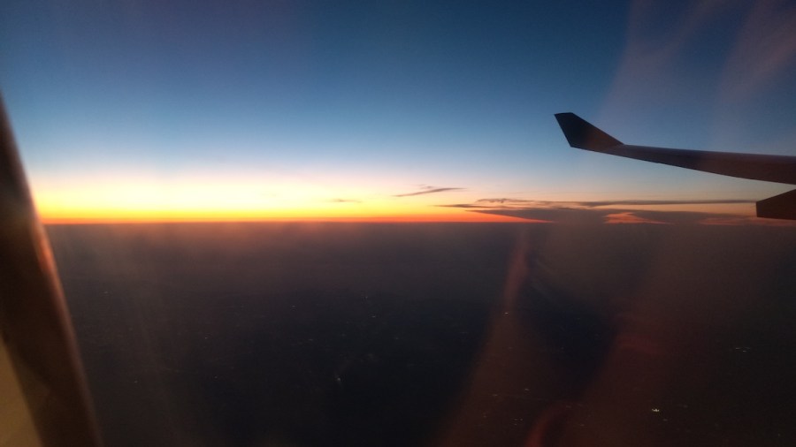 Photo shows the sun barely over the horizon, a jet plane wing visible in silhouette to the right. Taken somewhere over New York or the Atlantic Ocean.